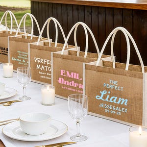 Pu&ograve; includere: Diverse borse di juta con manici bianchi sono disposte su un tavolo. Ogni borsa presenta un testo personalizzato in vari colori. Il tavolo &egrave; apparecchiato con piatti bianchi, posate dorate e candele accese, suggerendo un matrimonio o un evento.