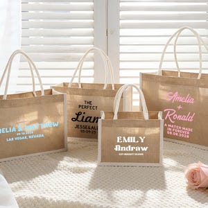 Pu&ograve; includere: Quattro borse tote in iuta con manici e finiture bianche. Ogni borsa presenta testo personalizzato in vari colori, inclusi nomi e date. Le borse sono disposte su una superficie bianca, con due rose rosa in primo piano.
