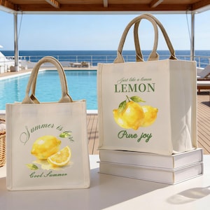 May include: Two canvas tote bags with beige handles and lemon illustrations. One bag reads "Summer is here" and "Cool Summer". The other bag reads "Just like a lemon LEMON Pure joy". The bags are on a white surface with a pool and ocean in the background.