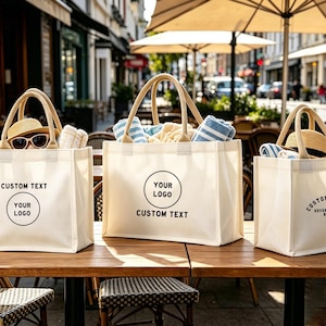 May include: Three cream tote bags with tan handles on a wooden table. Each bag has a circular logo area and the words "CUSTOM TEXT" printed on the front. The bags are filled with beach towels, sunglasses, and hats, perfect for a day out.