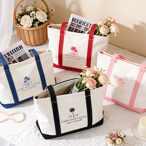 May include: Four white canvas tote bags with coloured handles and bases in red, blue, black, and pink. Each bag features a palm tree graphic, the text "2026", "CABO SAN LUCAS", and "amelia's bachelorette" printed on the front in a matching colour.