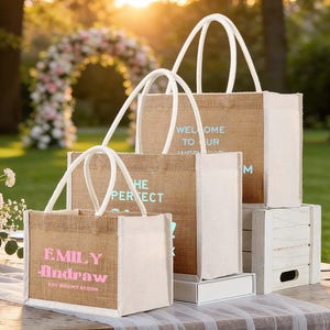 Pu&ograve; includere: Una collezione di borse tote in iuta con manici e finiture bianche, personalizzate con testo in vari colori. Le borse sono disposte su un tavolo, con un arco floreale e verde sullo sfondo. Il testo sulle borse include "Welcome to our wedding" e nomi.