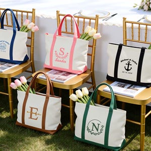 Puede incluir: Cinco bolsos de lona blancos con asas y bases de colores en azul, rosa, negro, marrón y verde. Cada bolso presenta un monograma o diseño personalizado con texto que incluye nombres y fechas, y contiene un ramo de tulipanes en su interior.