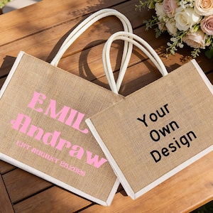 Pu&ograve; includere: Due borse tote in iuta con finiture e manici bianchi poggiano su una superficie di legno. Una borsa ha il testo "EMIL Andrew" in rosa, con testo aggiuntivo sottostante. L'altra borsa ha il testo "Your Own Design" in nero.