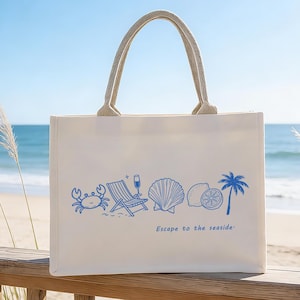Könnte beinhalten: Cremefarbene Tragetasche mit blauen Illustrationen zum Thema Meer, darunter eine Krabbe, ein Strandstuhl, eine Muschel, eine Zitrone und eine Palme. Sie hat beige Seilgriffe und den Text "Escape to the seaside".