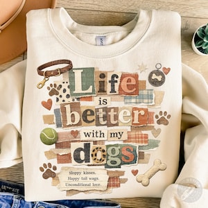 May include: Cream-colored sweatshirt with the text "Life is better with my dogs" in a collage style. The design includes dog paw prints, a tennis ball, hearts, and a dog bone. A dog collar and tag are also featured.