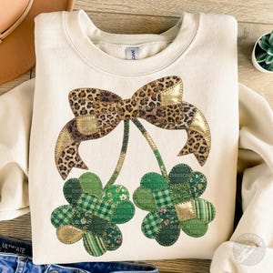 Può includere: Felpa color crema con un grande fiocco con stampa leopardata e due trifogli. I trifogli sono patchwork con motivi a quadri verdi, a righe e floreali. Sono visibili dettagli dorati.