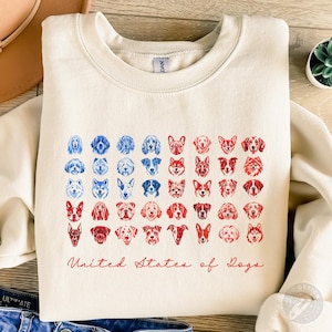 Op de afbeelding: Crèmekleurige sweatshirt met een afbeelding van honden gezichten in blauw en rood, gerangschikt als een vlag. De tekst "United States of Dogs" staat in rode letters. Een casual, comfortabel kledingstuk.