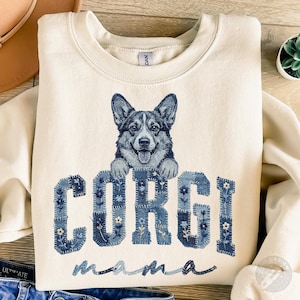 May include: Cream-colored sweatshirt featuring a smiling Corgi dog illustration above the word "CORGI" in denim patchwork letters. Below, the word "mama" is written in a cursive font. The design is centered on the front of the shirt.