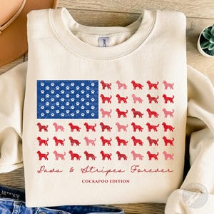 May include: A cream-colored crewneck sweatshirt featuring a graphic design of a flag composed of blue paw prints and rows of red and pink Cockapoo dog silhouettes. The text Paws & Stripes Forever and COCKAPOO EDITION is printed in red below.