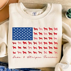 Op de afbeelding: Crèmekleurige sweatshirt met een patriottisch ontwerp. Het heeft een blauwe vlag met pootafdrukken en rode en roze honden silhouetten gerangschikt als strepen. De tekst "Paws & Stripes Forever" is in rood script geschreven.