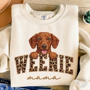 Può includere: Felpa color crema con un disegno di bassotto marrone. La parola "WEENIE" è in lettere con stampa leopardata, sopra la parola "mama" in corsivo. Il cane sorride, mostrando la lingua.