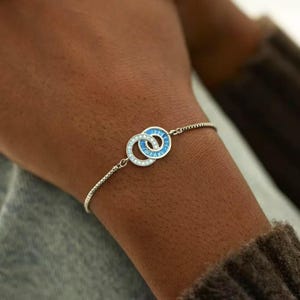 May include: Silver bracelet with two intertwined circles, one with blue baguette-cut stones and the other with clear stones. The bracelet has a fine chain and is displayed on a wrist.