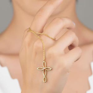 May include: A gold-colored necklace featuring a cross pendant with an infinity symbol at the bottom. The cross has four curved petals and a small, round, red gemstone in the center. The necklace is held by a person.