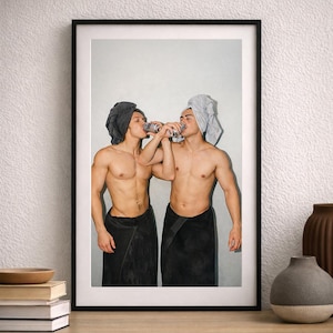 May include: Framed print featuring two individuals, each with a towel wrapped around their head and waist, drinking from cans. The image has a soft, neutral color palette, with a focus on the figures and their interaction. The print is displayed on a wooden surface.