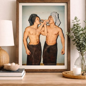 May include: Framed print featuring two individuals, each wearing a towel on their head and wrapped around their waist, drinking from cans. The image is set against a light blue background, with a wooden frame.