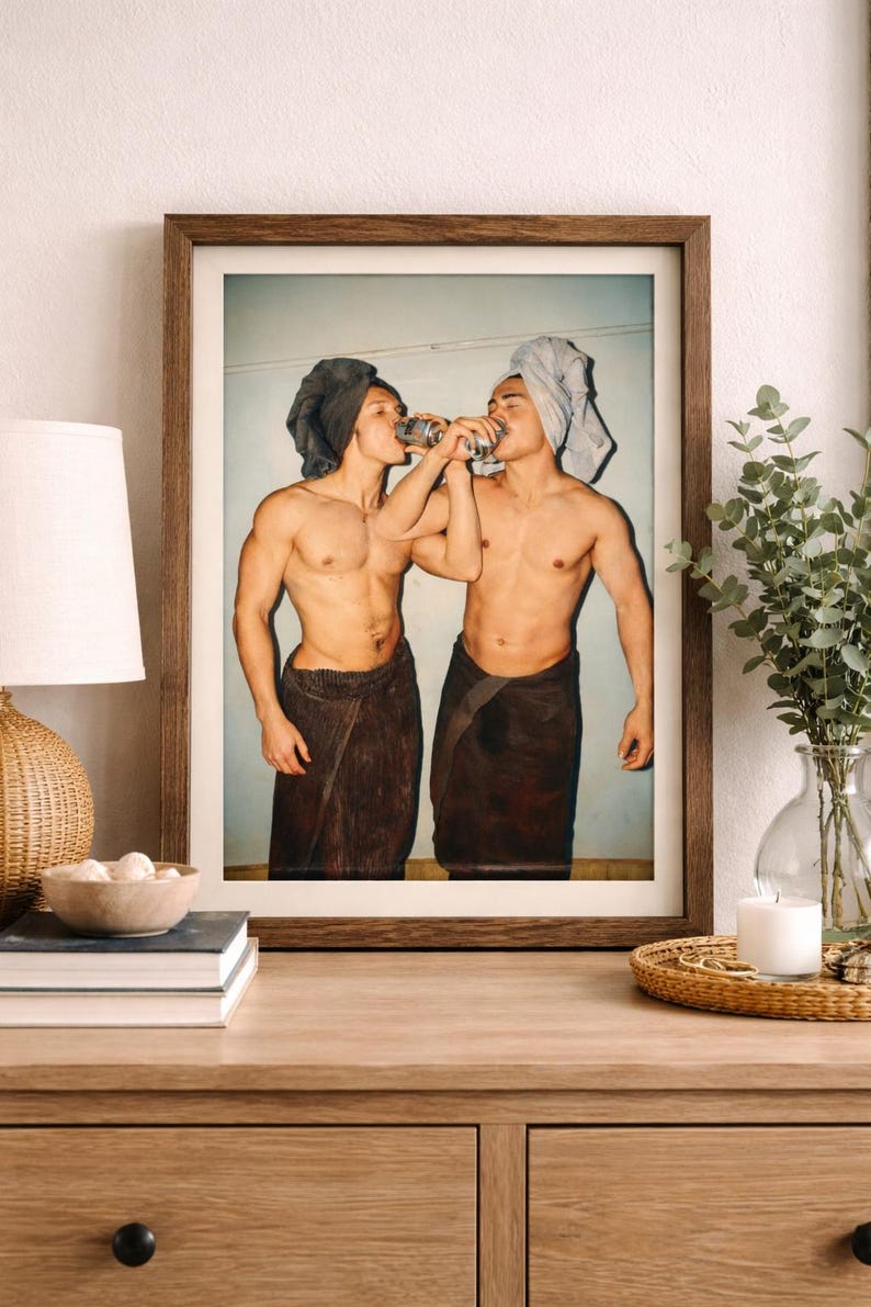 May include: Framed print featuring two individuals, each wearing a towel on their head and wrapped around their waist, drinking from cans. The image is set against a light blue background, with a wooden frame.