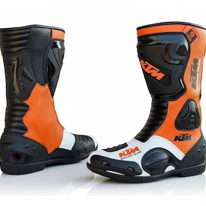 Könnte beinhalten: Paar orange, schwarze und weiße Motorradstiefel. Die Stiefel tragen das KTM Racing Team Logo und Schutzplatten. Sie sind für Sicherheit und Leistung konzipiert und haben ein hohes Design und eine verstärkte Konstruktion.
