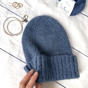 Puede incluir: Un gorro azul tejido con un dobladillo, junto a una madeja de lana azul con una etiqueta "Merino". También son visibles anillos dorados y un collar de cuentas con un dije azul.