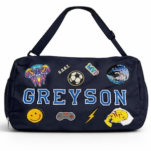 Puede incluir: Bolsa de deporte azul marino con correa para el hombro, decorada con parches que incluyen una calavera, un balón de fútbol y una carita sonriente. El nombre "GREYSON" se muestra en letras grandes y azules.