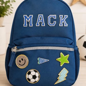 May include: A navy blue backpack featuring the name MACK in white and blue varsity-style letters. The front pocket is decorated with five patches: a smiley face, a blue pennant with the text LET'S GO!, a green star, a soccer ball, and a lightning bolt.