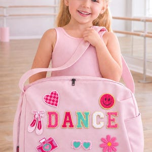 Op de afbeelding: Een roze danstas met patches, waaronder een hart, balletschoenen, een camera en een bloem. Het woord "DANCE" is in kleurrijke letters gespeld. Een jong meisje in een roze turnpakje draagt de tas.