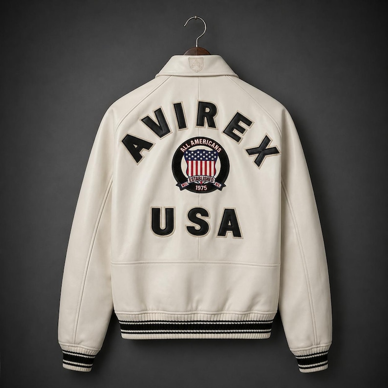May include: Cream-colored leather jacket with the word "AVIREX" across the back, along with "USA" and a circular patch featuring the American flag. The jacket has black and white striped cuffs and hem.