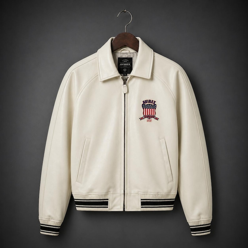 May include: Cream-colored leather jacket with a zip-up front and a collared neckline. Features a patch on the chest with a red, white, and blue emblem. The jacket has black and white striped cuffs and hem. The brand name "AVIREX" is visible.