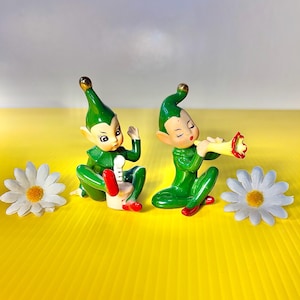 May include: Two vintage ceramic elf figurines in green outfits with red shoes. One elf plays a musical instrument, the other gestures. Daisies with white petals and yellow centers are on either side. The background is yellow.