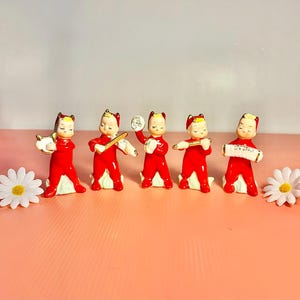 May include: A collection of five vintage ceramic figurines dressed in red outfits, each playing a different musical instrument. The figurines have a white base and gold accents. Two white daisies are placed on either side of the figurines.