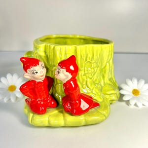 May include: A vintage ceramic planter in the shape of a tree stump, painted in bright yellow-green. Two red elf figurines are positioned in front of the planter. The word "Love" is embossed on the side. White daisy flowers are on either side.