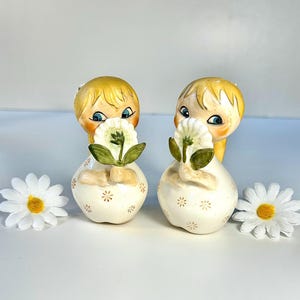 May include: Two vintage ceramic figurines of girls holding flowers. The figurines have blonde hair, blue eyes, and are dressed in white dresses with gold star accents. Two white daisies with yellow centers are on either side.