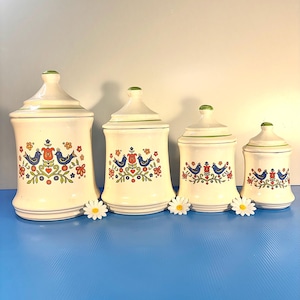 May include: Set of four off-white ceramic canisters with lids. Each canister features a colorful floral and bird design in red, blue, and green. The lids have a green trim and finial. A decorative kitchen storage set.