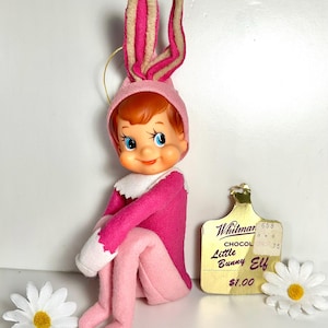 May include: A vintage Little Bunny Elf doll wearing a pink outfit and bunny ears. The doll has a cheerful expression and is sitting down. A gold tag reads "Whitman Chocolate Little Bunny Elf". White daisy flowers are placed near the doll.