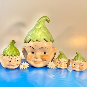 May include: A collection of vintage ceramic elf-like planters with green leaf-shaped hats and smiling faces. The planters vary in size, with the largest one in the center and smaller ones on either side. White daisy flowers are placed in front of the planters.