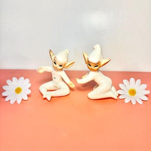 May include: Two ceramic elf figurines with pointed ears and hats, posed on a pink surface. The elves are white with tan accents and dark eyes. White daisy flowers with yellow centers are placed on either side of the figurines.