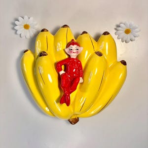 May include: A vintage ceramic banana-shaped wall pocket with a smiling red elf figurine. The bananas are bright yellow with brown tips. Two white daisies with yellow centers are on either side of the banana wall pocket.