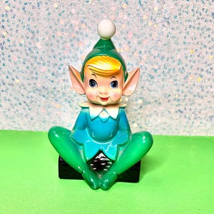 May include: A ceramic elf figurine featuring a green hat with a white pom-pom, blonde hair, and large blue eyes. The elf wears a teal tunic with a white collar and sits with legs crossed on a black base, displaying pointed ears and a smiling face.