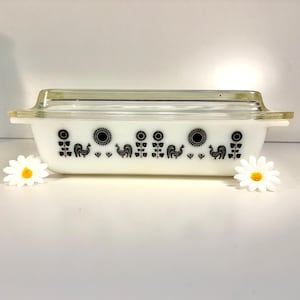 May include: A rectangular white casserole dish with a clear glass lid. The dish features a black rooster, sunflower, and floral pattern. The dish is a vintage kitchenware item.