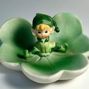 May include: A ceramic figurine of a seated elf with blonde hair and a green outfit, resting in a green, clover-shaped dish. The figurine has a serene expression and is painted with delicate details. The dish has a white rim.