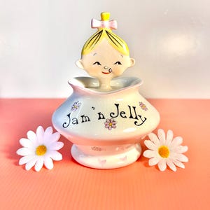 May include: A vintage ceramic jam and jelly jar with a smiling girl figurine on top. The jar is white with pink accents and the words "Jam 'n Jelly" in black script. Decorative daisies are painted on the jar. Two white daisies with yellow centers are placed on either side.
