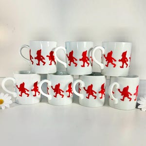 May include: A collection of eight white ceramic mugs. Each mug features a red graphic of a marching gnome. The mugs are arranged in a stacked display, showcasing their design and shape.