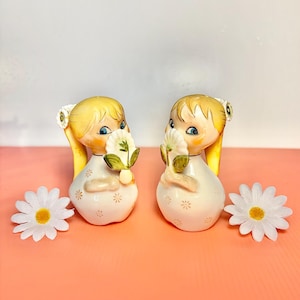 May include: Two vintage ceramic figurines of girls with blonde pigtails, holding white flowers. The figurines are white with floral details and are set on a pink surface. Two white daisies with yellow centers are also present.