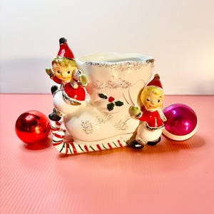 May include: A vintage Christmas boot-shaped planter with two elf figurines. The white ceramic boot is decorated with gold glitter and holly. Two red and pink Christmas ornaments sit nearby. The elves wear red and white outfits.
