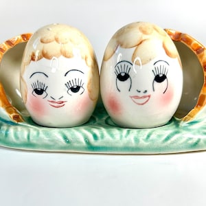 May include: A vintage salt and pepper shaker set featuring two egg-shaped figures with painted faces, nestled in a shell-like holder. The base is light green with a leaf design. The faces have rosy cheeks, eyelashes, and smiling lips. Two white daisies are on either side.