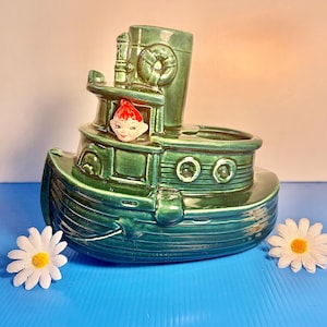 May include: A vintage green ceramic tugboat planter with a smiling face figure in the window. The boat has a smokestack, a wheel, and decorative details. Two white daisies with yellow centers are placed near the boat on a blue surface.