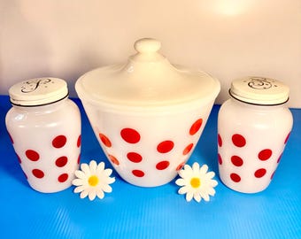 May include: A vintage white milk glass kitchen set with red polka dots. The set includes a lidded jar and two shakers with metal lids. The jar has a rounded shape, and the shakers are cylindrical. Decorative daisy accents are placed in front of the set.