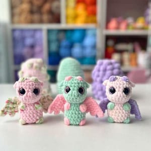 May include: Three handmade crochet dragon toys in pink, green, and lavender. Each dragon has large black eyes and small wings. The dragons are displayed on a white surface with a blurred background of colourful yarn.