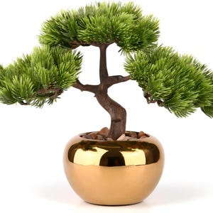May include: A decorative bonsai tree in a gold-colored, round pot. The tree has green, textured foliage and a brown trunk. Small pebbles are visible at the base of the tree. The pot has a shiny, reflective surface.