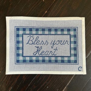 May include: A rectangular needlepoint canvas with the phrase "Bless your Heart" in cursive script. The canvas features a blue and white plaid border and a white background. A small "C" is in the lower right corner.